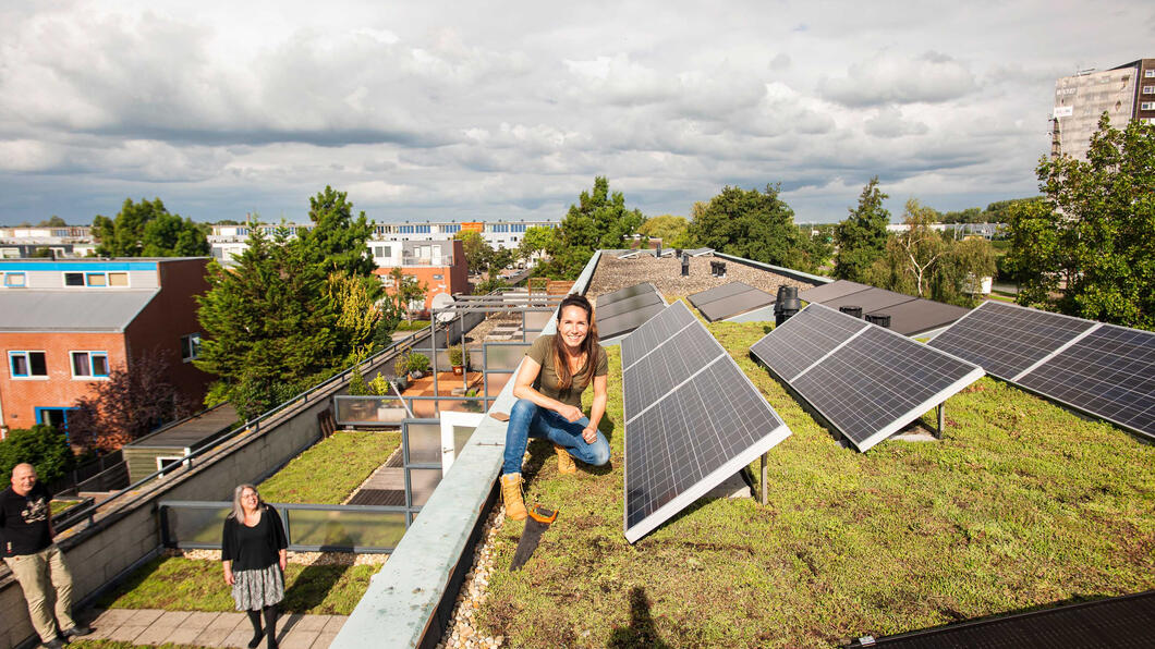 Een woonwijk met groene daken en zonnepanelen. Een vrouw zit naast een zonnepaneel en lacht naar de camera.
