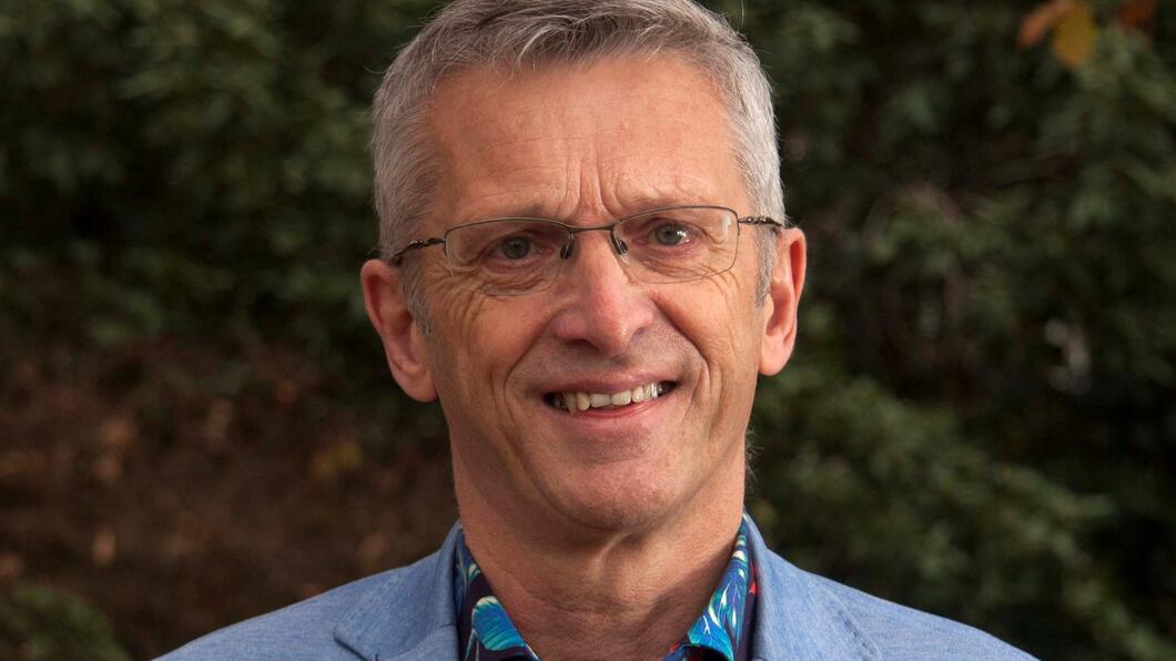 Henk Nijmeijer. Hij is een witte man met grijs haar. Henk heeft een blauw jasje aan en een bont shirt eronder. Hij lacht naar de camera.