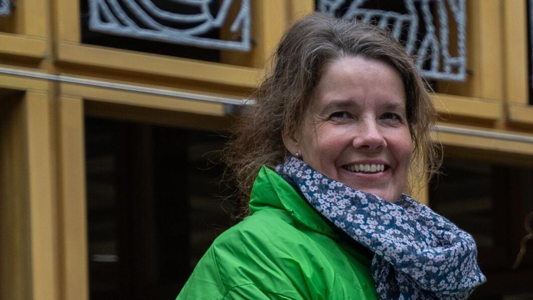 Verone Meengs - de Vries heeft een groene jas aan en een blauwe sjaal om. Achter haar staat een wand met kunstzinnig vormgegeven ruiten.