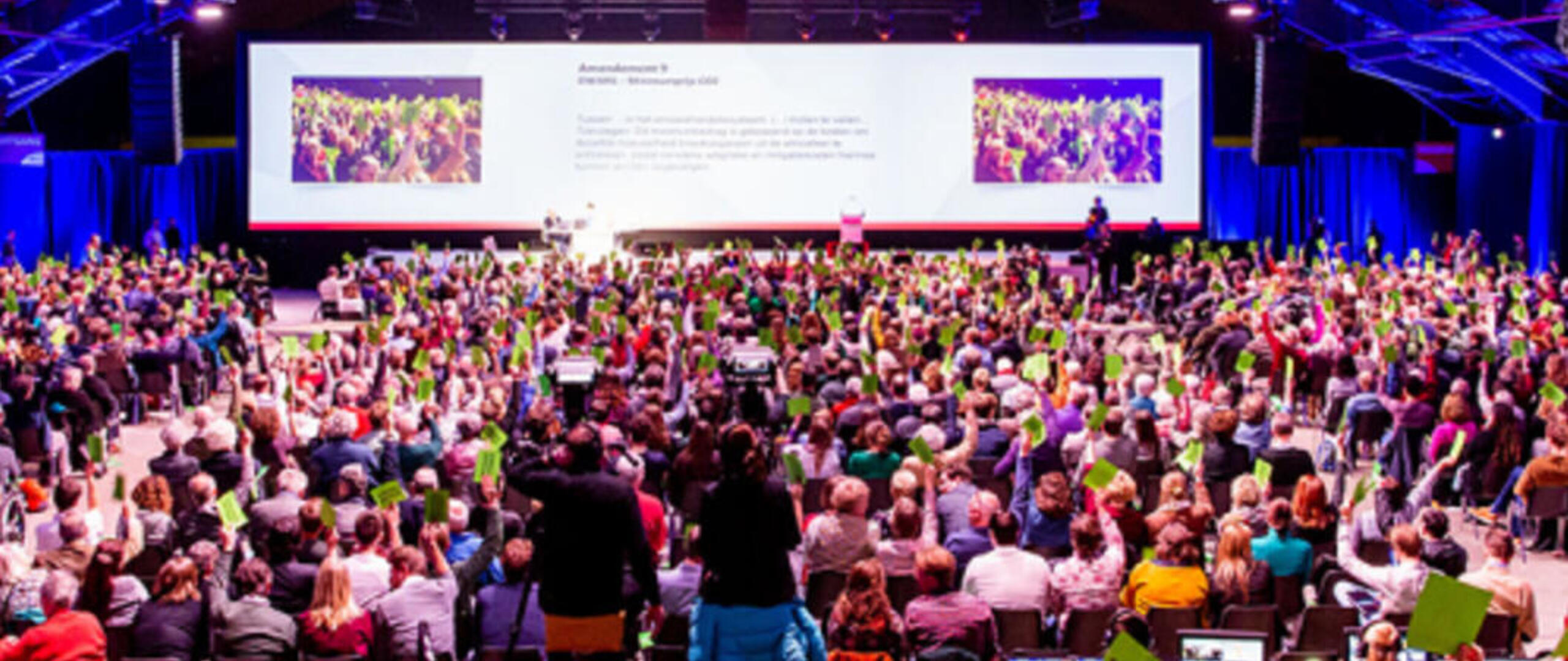 Een GroenLinks-congres, foto genomen vanuit achterin de zaal. Er zitten veel mensen in de zaal, vooraan hangen grote schermen.