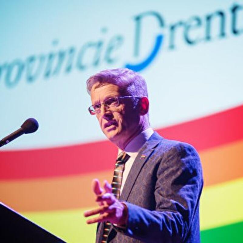 Henk Nijmeijer spreekt op een podium. Achter hem is een regenboogvlag in beeld en 'provincie Drenthe'
