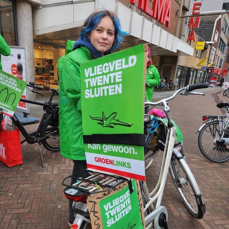 Een tiener staat bij een fiets met daarop een bord 'vliegveld Twente sluiten. Kan gewoon.'