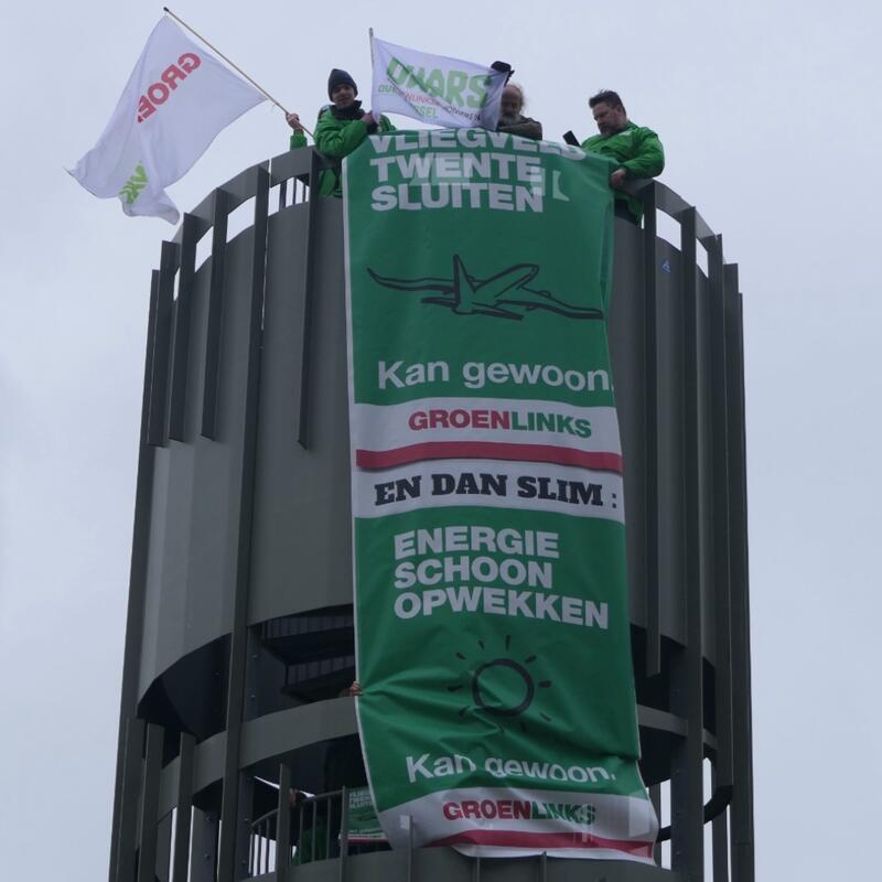 GroenLinksers boven op de toren op vliegveld Twente. Er hangt een groot spandoek met daarop de tekst "Vliegveld twente sluiten en dan slim schone energie opwekken. Kan gewoon.'
